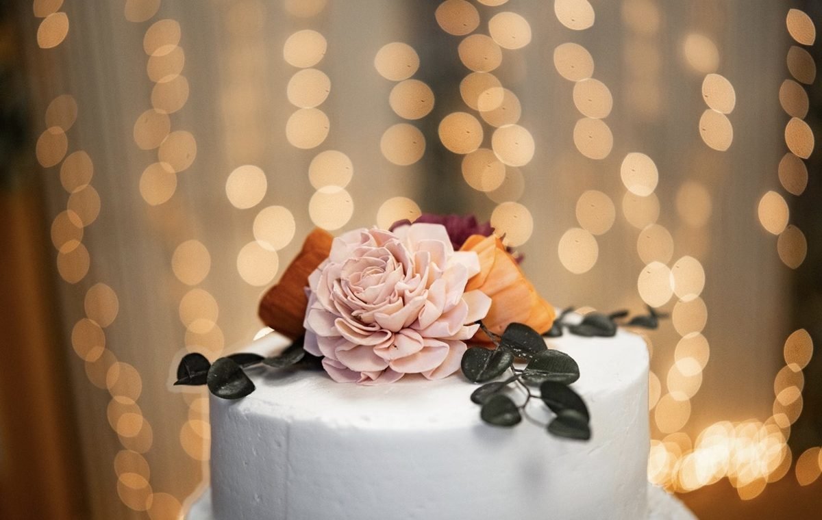 A white frosted cake topped with flowers, against warm string lights
