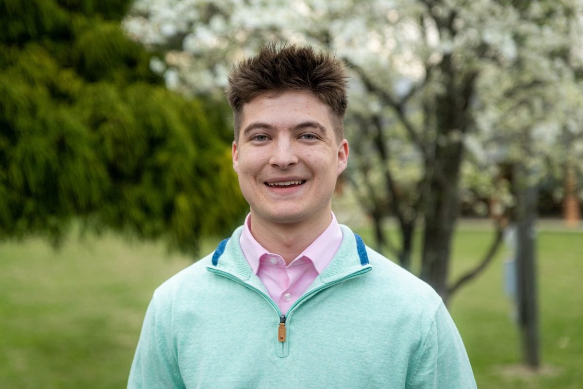 A smiling young man in a mint quarter-zip, with a blossoming tree behind him