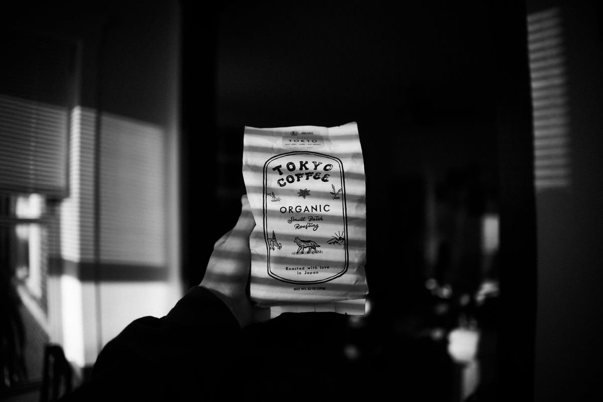 In black and white, a hand holds up the bag in a dark room, striped with light from the blinds