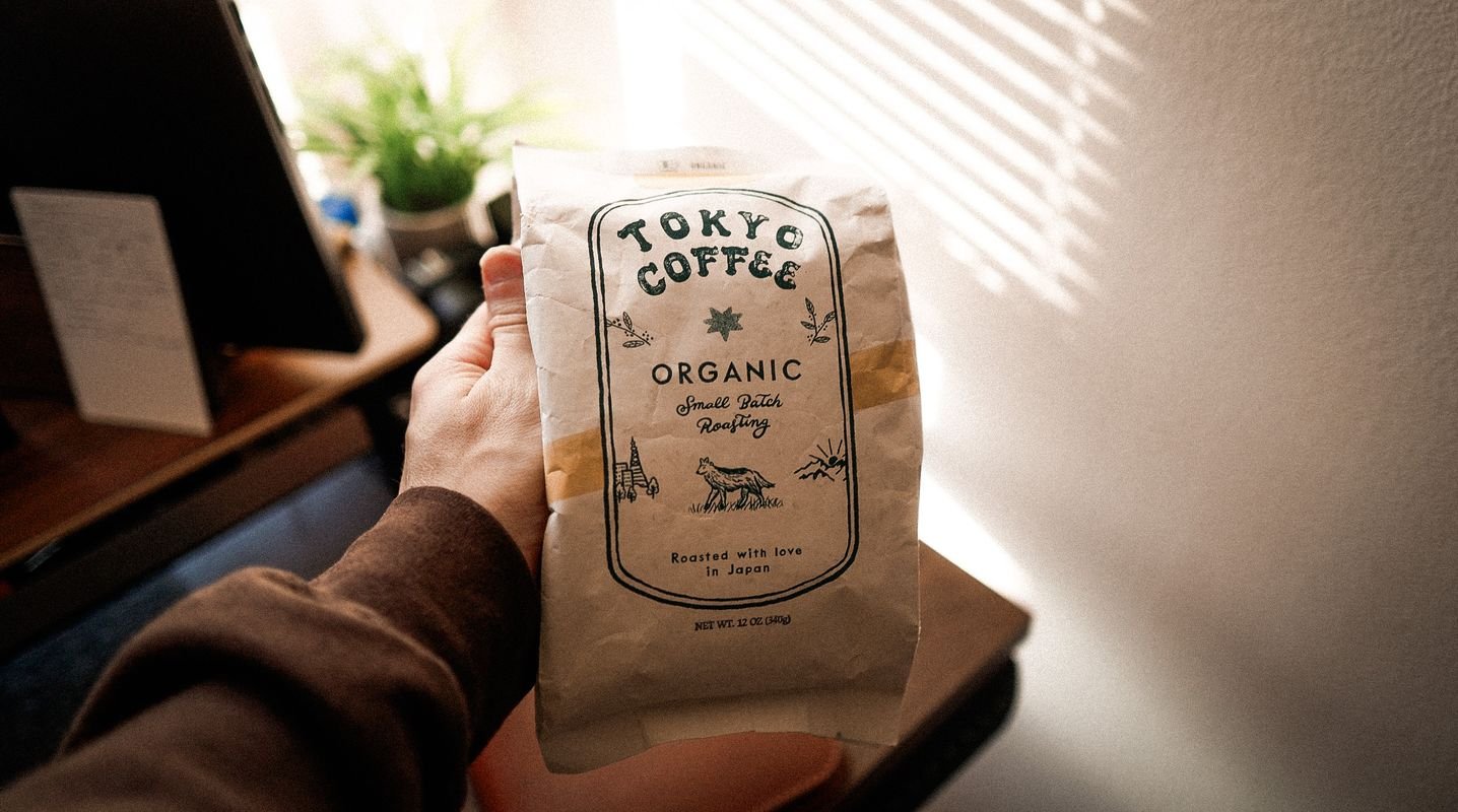 A hand holding the Tokyo Coffee bag beside a sunny window and a plant