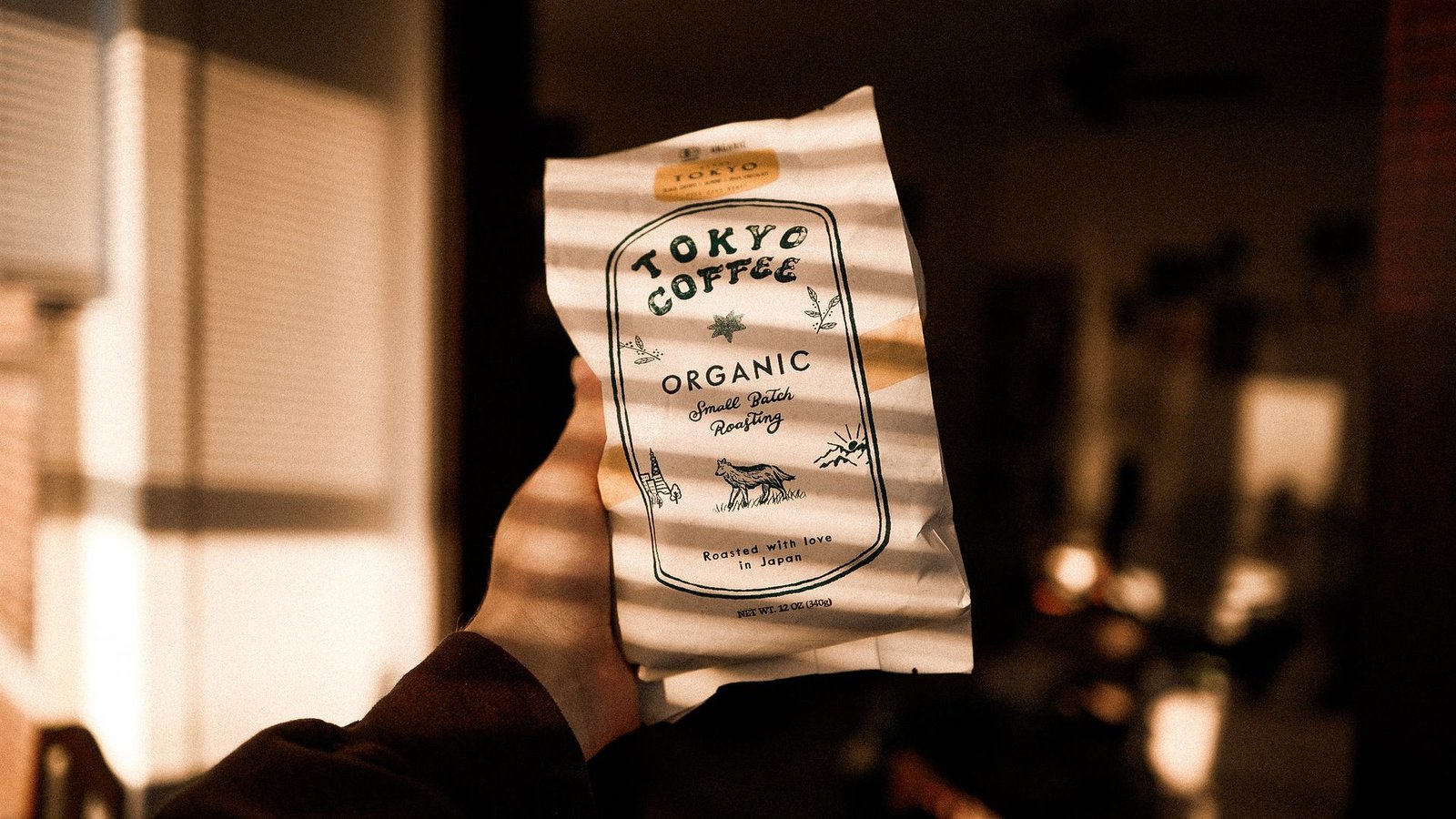A hand holding up a bag of Tokyo Coffee, striped with light through window blinds