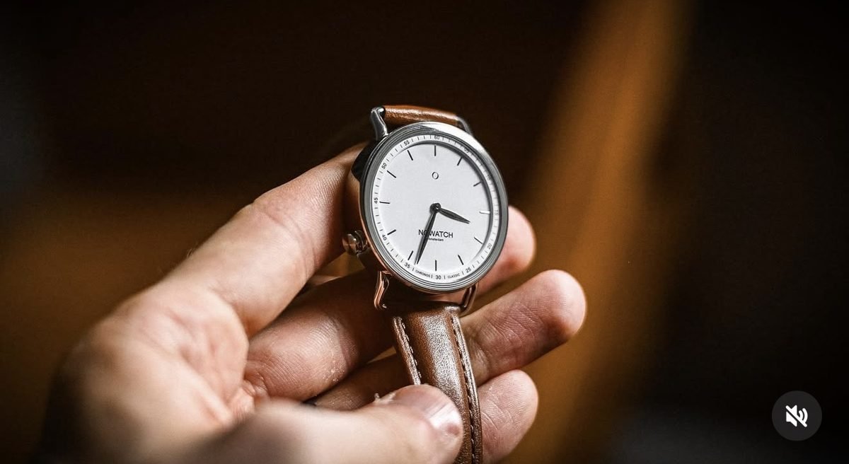 A hand holding a watch with a white face and tan leather strap
